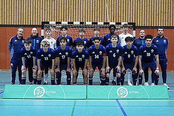 Das Bild zeigt eine Futsal-Mannschaft in einer Sporthalle. Die Gruppe steht in zwei Reihen vor einem Handballtor. Alle tragen dunkelblaue Trikots mit weißen Nummern und schwarzen Shorts. Vor der Mannschaft liegen zwei grüne Banden mit dem DFB-Logo und der Aufschrift „FUTSAL“. Der Hintergrund besteht aus einer Holzvertäfelung und dem Tor.