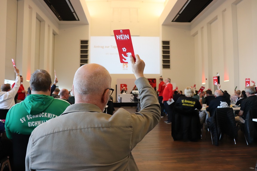 "In einem großen Raum mit hohen Decken und Fenstern hebt ein Mann eine rote Karte mit der Aufschrift 'NEIN' hoch. Weitere Personen im Raum halten ebenfalls rote Karten. Einige tragen Kleidung mit Vereinsnamen wie 'SV Schmöckwitz Eichwalde'. Im Hintergrund ist eine Leinwand mit unlesbarem Text zu sehen."