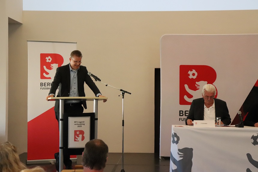 Ein Mann spricht an einem Rednerpult, während ein anderer Mann an einem Tisch sitzt. Im Hintergrund sind Banner des Berliner Fußball-Verbands und der BFV-Jugend zu sehen. Menschen im Vordergrund hören zu. Der Redner am Pult ist Tobias Dollase.