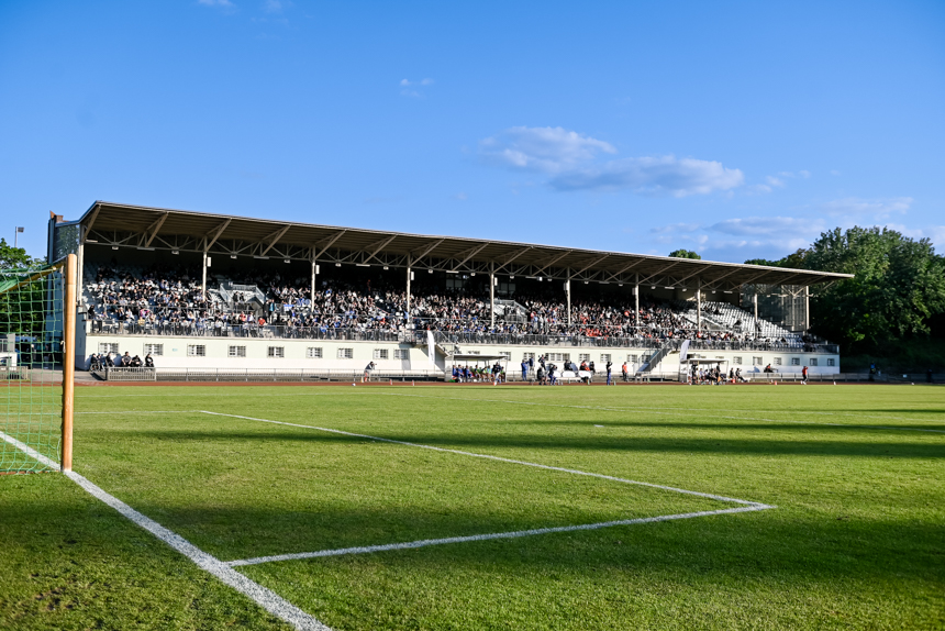 Das Poststadion mit überdachter Tribüne ist gut gefüllt mit zahlreichen Zuschauerinnen und Zuschauern. Der Rasen auf dem Spielfeld ist gepflegt und leuchtend grün. Auf der linken Seite des Bildes ist ein Tor zu sehen. Einige Personen stehen oder gehen am Spielfeldrand entlang. Der Himmel ist größtenteils klar mit wenigen Wolken, was auf gutes Wetter hinweist. Foto: Sandra Ritschel