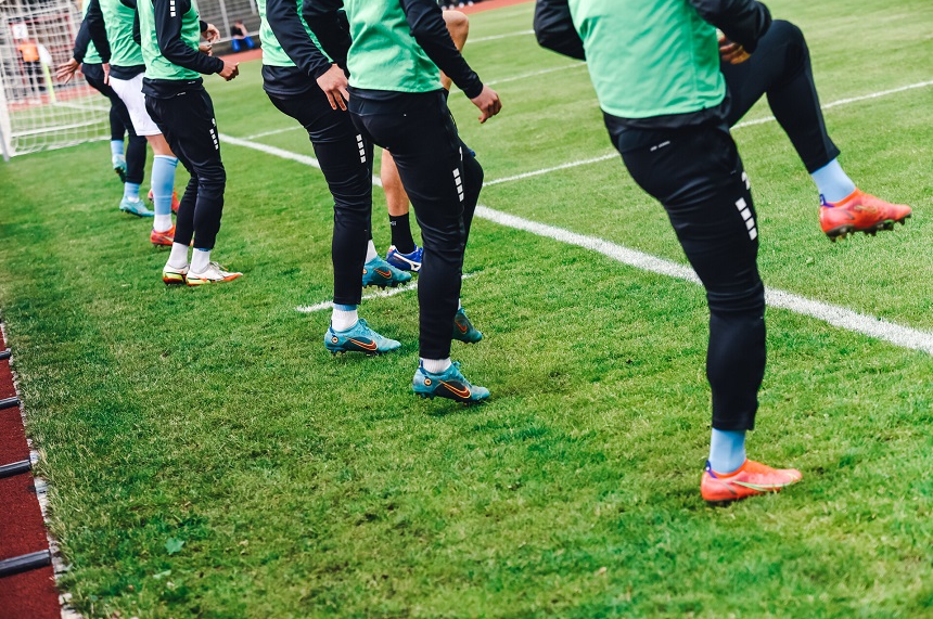 Auf einem Fußballfeld wärmt sich eine Gruppe von Spielern auf. Sie tragen grüne Trainingswesten, schwarze Hosen und bunte Fußballschuhe. Einige führen Dehnübungen aus, indem sie ihre Beine heben. Im Hintergrund ist ein Fußballtor sichtbar.