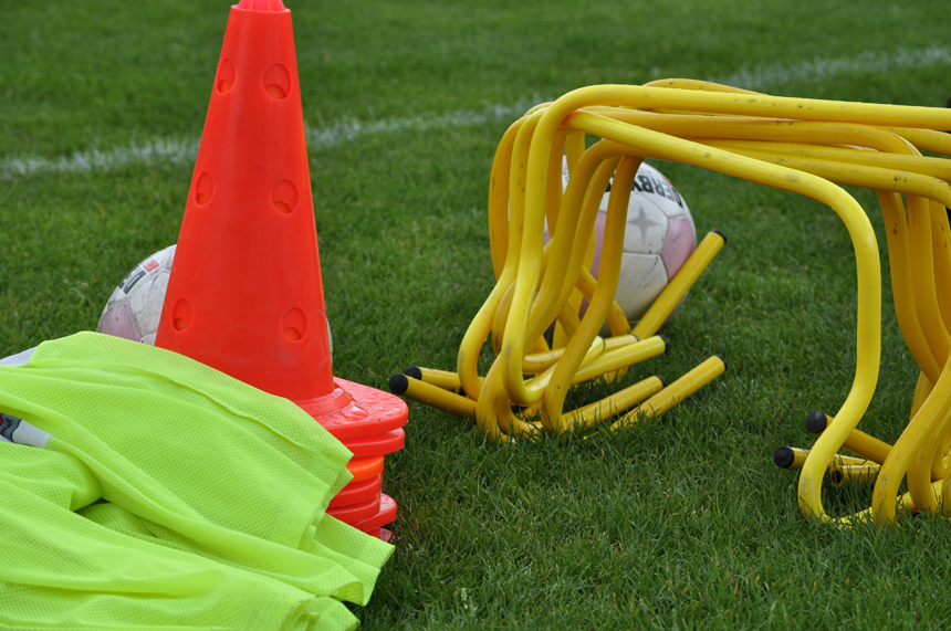 Auf einer grünen Rasenfläche liegt eine Auswahl an Trainingsmaterialien: orangefarbene Hütchen, gelbe Koordinationshürden, neongrüne Leibchen und zwei Fußbälle.