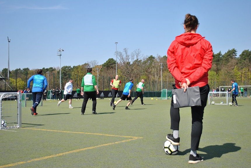 Eine Gruppe von Menschen spielt Fußball auf einem Kunstrasenplatz im Freien. Im Vordergrund steht eine Person in einer roten Jacke mit einem Klemmbrett und einem Fußball unter dem Fuß, die das Spiel beobachtet. Im Hintergrund sind mehrere Spieler in verschiedenen farbigen Trikots zu sehen, die dem Ball nachjagen. Der Platz ist von Bäumen umgeben und der Himmel ist klar und blau. Foto: BFV