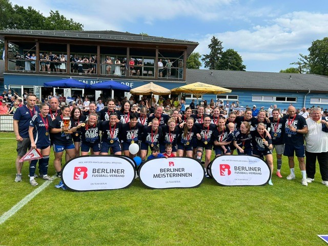 Auf einem Fußballfeld steht die Frauenmannschaft von B.W. Hohen Neuendorf. Die Spielerinnen tragen Medaillen um den Hals und posieren gemeinsam für ein Foto. Vor der Gruppe stehen drei Schilder mit der Aufschrift „BERLINER FUSSBALL-VERBAND“ und „BERLINER MEISTER“. Im Hintergrund sind Bäume und ein klarer Himmel zu sehen.