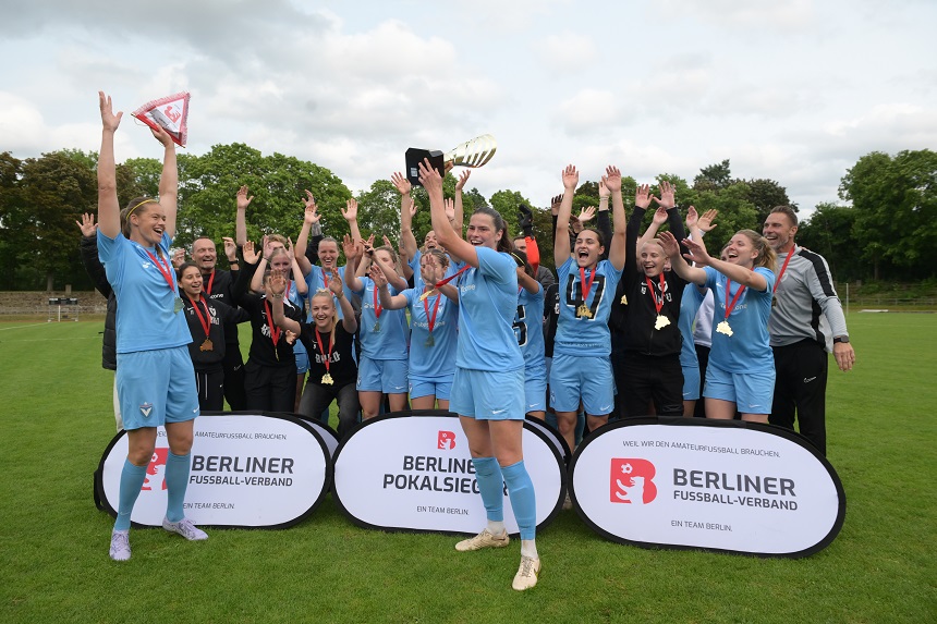 Eine Gruppe von Fußballspielern und Betreuern feiert auf einem grünen Rasenplatz einen Sieg. Die Spieler tragen hellblaue Trikots, einige halten einen Pokal in die Höhe. Vor der Gruppe stehen drei Banner mit der Aufschrift „BERLINER FUSSBALL-VERBAND“ und „BERLINER POKALSIEGER“. Im Hintergrund sind Bäume und ein bewölkter Himmel zu sehen.