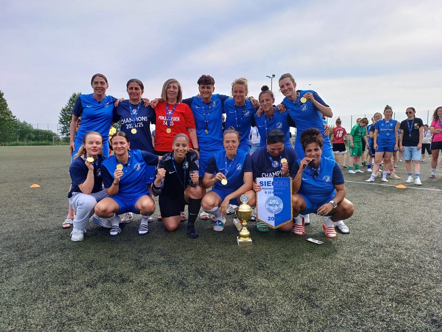 Auf einem Fußballfeld steht eine Gruppe von Frauen in blauen Trikots. Sie tragen Medaillen und halten Pokale in den Händen – ein Hinweis auf einen gewonnenen Titel. Auf den Trikots steht „CHAMPIONS 2024/25“. Im Vordergrund ist ein Banner mit der Aufschrift „SIEGER“ und der Jahreszahl „2025“ zu sehen.