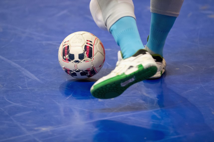 Das Foto zeigt eine Nahaufnahme eines Fußballspielers während eines Spiels oder Trainings in einer Halle. Im Mittelpunkt steht ein Fußball, der gerade vom Spieler kontrolliert wird. Der Ball ist überwiegend weiß und mit roten sowie schwarzen Mustern versehen. Der Spieler trägt weiße Sportschuhe mit grüner Sohle, hellblaue Socken und weiße Sportkleidung. Der Boden ist aus glattem, blauem Hallenbelag. Weitere Personen oder Elemente sind auf dem Bild nicht sichtbar – der Fokus liegt ganz auf dem Moment der Ballkontrolle.