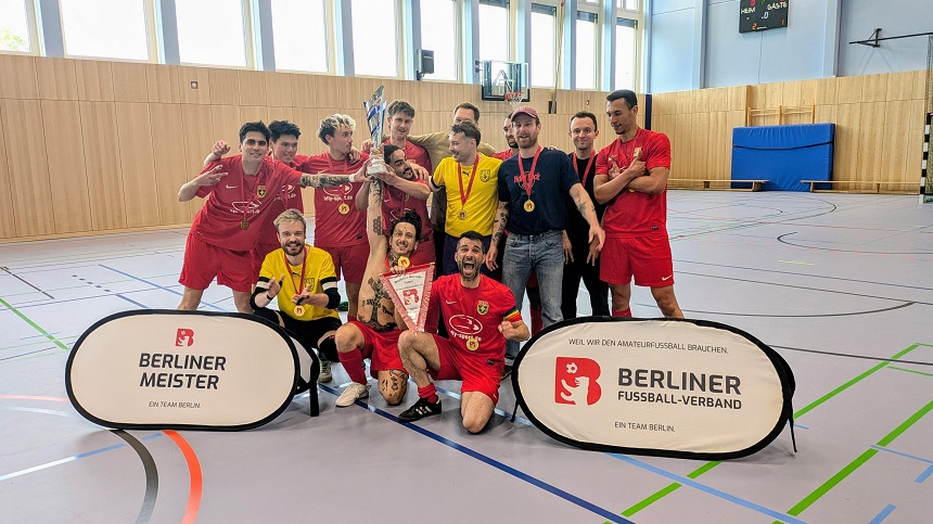 In einer Sporthalle steht eine Gruppe von Menschen in roten Trikots zusammen. Sie posieren für ein gemeinsames Foto. Einige von ihnen halten Medaillen und einen Pokal in den. Vor der Gruppe stehen zwei Schilder mit der Aufschrift „BERLINER MEISTER“ und „BERLINER FUSSBALL-VERBAND“.