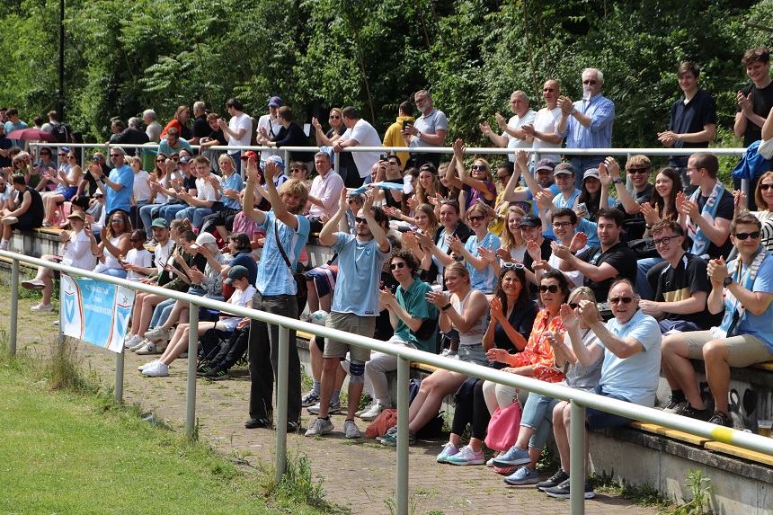 Das Bild zeigt eine größere Menschenmenge auf einer Tribüne im Freien. Einige Personen sitzen, andere stehen. Viele klatschen oder jubeln, was auf eine sportliche oder festliche Veranstaltung hinweist. Die Menschen tragen überwiegend legere Kleidung. Foto: BFV