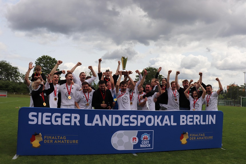 Mehrere Personen stehen jubelnd hinter einem großen blauen Banner mit der Aufschrift „SIEGER LANDESPOKAL BERLIN“. Auf dem Banner sind zusätzlich die Logos und Schriftzüge „FINALTAG DER AMATEURE“ und „COSY WASCH“ zu sehen. Die Personen tragen Medaillen um den Hals, eine Person hält einen Pokal in die Höhe. Die Szene vermittelt den Moment eines sportlichen Sieges.