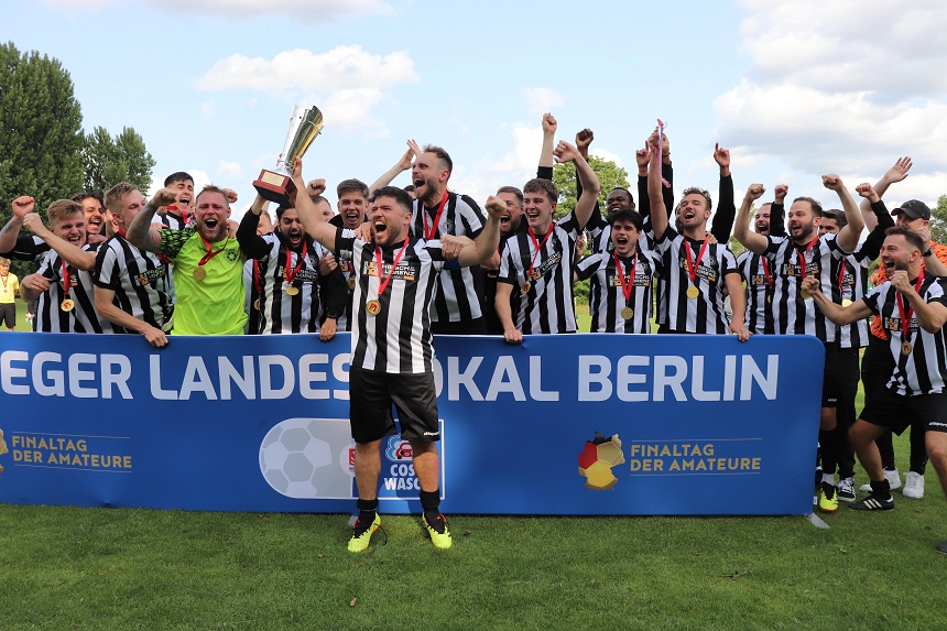 Eine Gruppe von Fußballspielern steht jubelnd hinter einem großen blauen Banner mit der Aufschrift „SIEGER LANDESPOKAL BERLIN“. Die Spieler tragen schwarz-weiß gestreifte Trikots. Einige von ihnen haben Medaillen um den Hals. Ein Spieler hält einen Pokal in die Höhe. Auf dem Banner sind außerdem die Logos und Schriftzüge „FINALTAG DER AMATEURE“ und „COSY WASCH“ zu sehen. Die Stimmung wirkt feierlich und siegreich.
