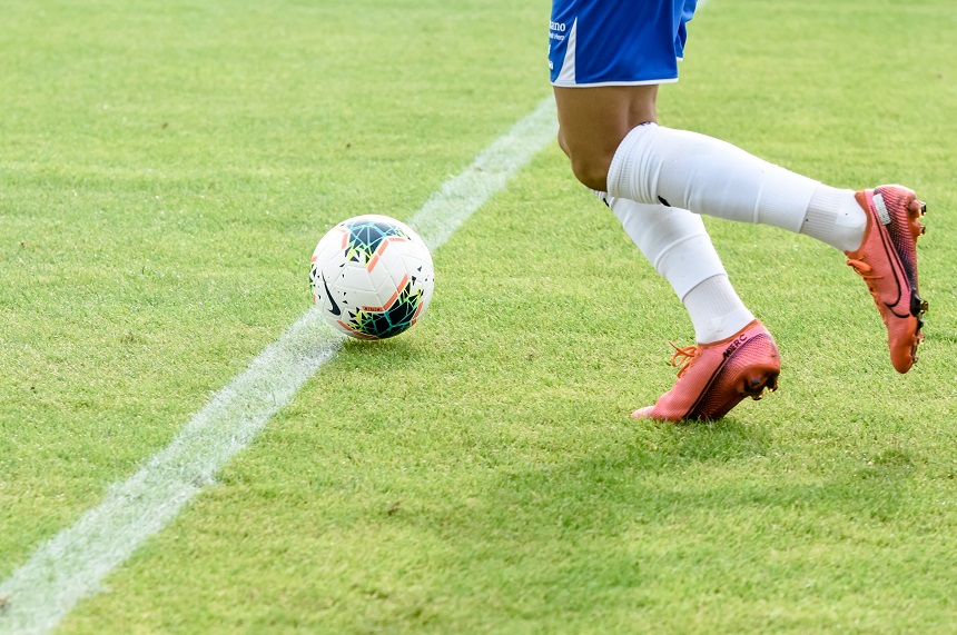 Das Foto zeigt eine Szene auf einem Fußballfeld. Im Vordergrund ist ein Fußballspieler zu sehen, der sich in Bewegung befindet. Er trägt weiße Socken, orangefarbene Fußballschuhe und eine blaue Sporthose. Der grüne Rasen ist gut gepflegt und wird von einer weißen Linie diagonal durchzogen, die das Spielfeld in zwei Bereiche unterteilt. Am linken Bildrand ist ein Fußball zu erkennen, der ebenfalls in Bewegung zu sein scheint. Die Perspektive des Fotos lässt vermuten, dass der Spieler dem Ball hinterherläuft oder ihn gleich erreichen wird.
