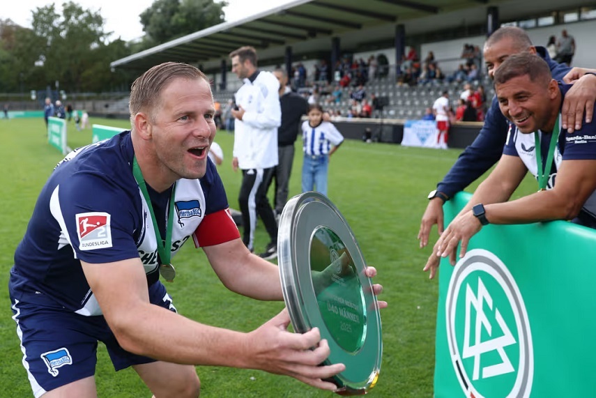 Das Foto zeigt eine Szene auf einem Fußballplatz nach einem Spiel. Im Vordergrund hält eine Person in einem dunkelblauen Trikot mit weißen Details eine große silberne Meisterschale in den Händen. Auf dem linken Ärmel des Trikots ist das Logo der Bundesliga zu sehen, und am Oberarm trägt die Person eine rote Kapitänsbinde. Um den Hals hängt eine grüne Medaille. Die Meisterschale ist rund und glänzend, mit einer Gravur in der Mitte, die den Titel „Deutscher Meister“ und das Jahr erkennen lässt. Rechts im Bild lehnt eine andere Person über eine grüne Bande mit dem DFB-Logo und streckt die Hände in Richtung der Schale, als würde sie diese berühren oder entgegennehmen. Im Hintergrund sind weitere Personen auf dem Spielfeld zu sehen, einige in Sportkleidung, andere in Freizeitkleidung. Dahinter befindet sich eine Tribüne mit Sitzplätzen, auf denen Zuschauer sitzen. Die Stimmung wirkt wie bei einer Siegerehrung oder einem feierlichen Moment nach einem gewonnenen Wettbewerb.