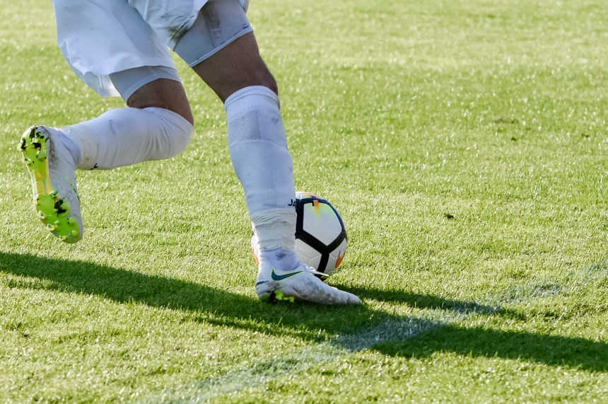 Das Foto zeigt eine Nahaufnahme von zwei Beinen einer Person auf einem grünen Rasen. Die Person trägt helle Sportschuhe mit farbigen Akzenten sowie lange, helle Sportsocken und kurze Sporthose. Ein Fußball liegt direkt vor einem der Füße und wird offenbar gerade geführt oder gestoppt. Der andere Fuß ist leicht angehoben, als befände sich die Person in Bewegung. Der Rasen ist gleichmäßig gemäht und von Sonnenlicht beschienen, dadurch entstehen klare Schatten auf dem Boden. Weitere Personen oder Objekte sind nicht zu sehen; der Bildausschnitt konzentriert sich ausschließlich auf die untere Körperhälfte, den Ball und die Bewegungssituation im Sportkontext.