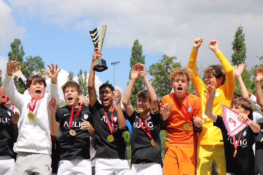 Eine Gruppe von Kindern feiert gemeinsam den Gewinn des Nike Youth Cups. Sie tragen Sportkleidung in verschiedenen Farben – darunter schwarze Trikots mit der Aufschrift „MULIG BAU“, sowie weiße und gelbe Outfits. Viele der Kinder haben Medaillen um den Hals. Der Pokal wird von mehreren Kindern in die Höhe gehalten. Die Gesichter der Kinder sind nicht erkennbar. Im Hintergrund sind Bäume und ein teilweise bewölkter Himmel zu sehen.