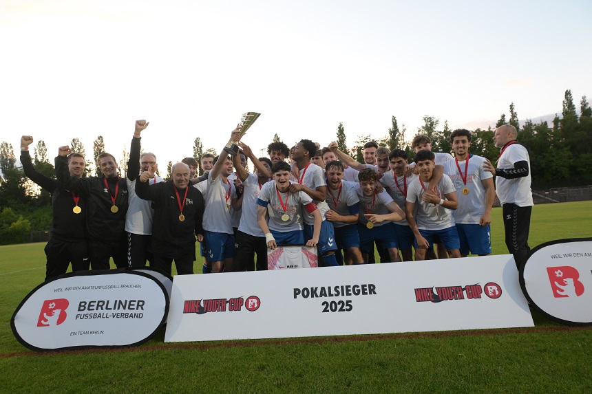 Eine Gruppe von Menschen steht auf einem Fußballfeld und feiert gemeinsam. Sie halten einen Pokal in die Höhe und tragen Medaillen um den Hals. Vor der Gruppe stehen zwei große Schilder:  Das linke Schild trägt die Aufschrift: „BERLINER FUSSBALL-VERBAND EIN TEAM BERLIN“. Das rechte Schild trägt die Aufschrift: „POKALSIEGER 2025“. Foto: Sandra Ritschel