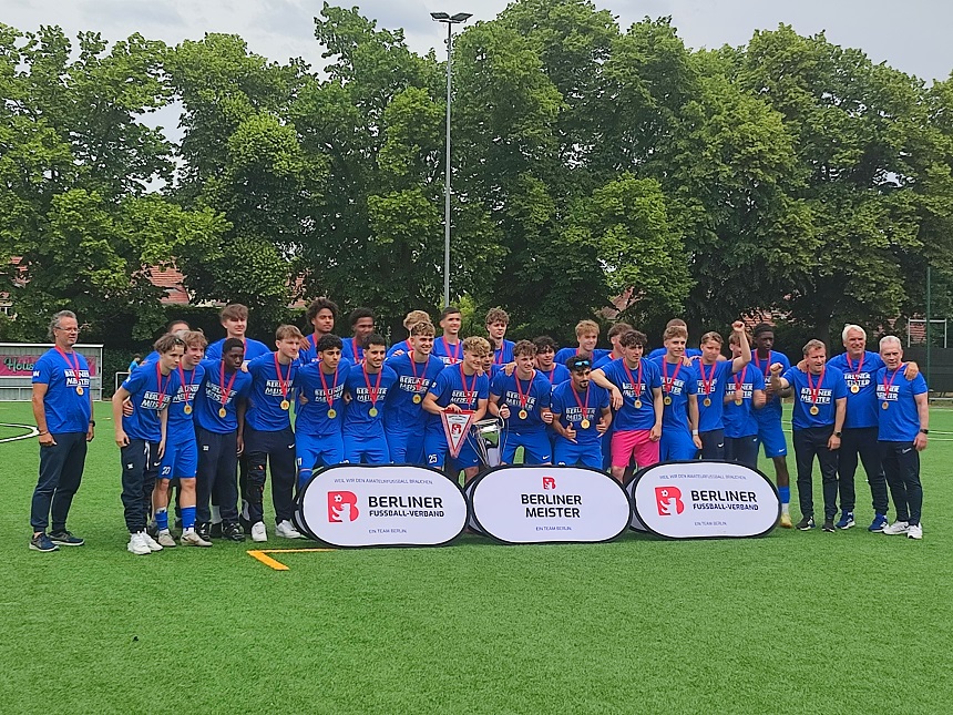 Auf einem Fußballfeld steht eine Gruppe junger Spieler und Trainer des SC Staaken. Alle tragen blaue Trikots und Medaillen um den Hals. In der Mitte hält ein Spieler einen Pokal. Vor der Gruppe stehen drei Schilder mit der Aufschrift „BERLINER FUSSBALL-VERBAND“, „BERLINER MEISTER“ und erneut „BERLINER FUSSBALL-VERBAND“. Im Hintergrund sind Bäume und ein weiteres Spielfeld zu sehen.