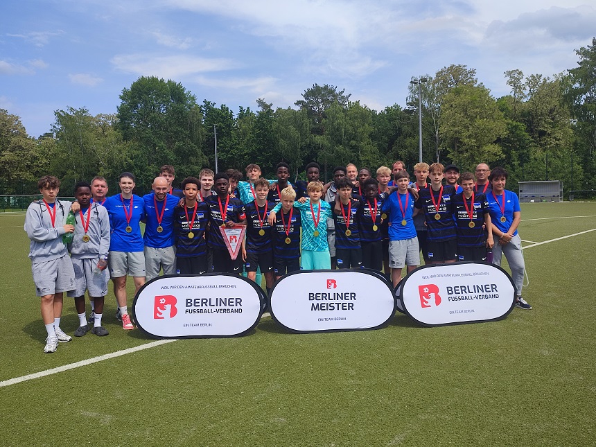 Auf einem Fußballfeld steht eine Gruppe junger Sportler:innen in Sportkleidung. Sie tragen Medaillen um den Hals und posieren für ein gemeinsames Foto. Vor der Gruppe stehen drei Schilder mit der Aufschrift „BERLINER FUSSBALL-VERBAND“ und „BERLINER MEISTER“. Im Hintergrund sind Bäume und ein klarer Himmel zu sehen.
