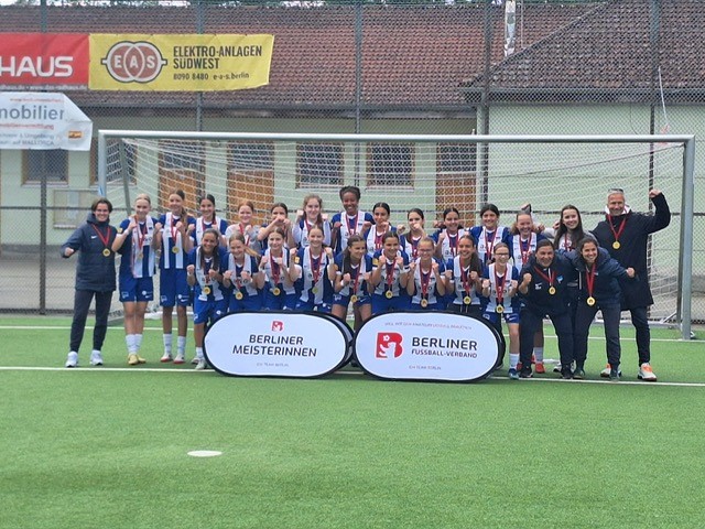 Auf einem Fußballfeld steht eine Gruppe von Personen, vermutlich ein Frauenteam, vor einem Tor und posiert für ein gemeinsames Foto. Die Teammitglieder tragen Medaillen. Vor der Gruppe liegen zwei Banner mit der Aufschrift „BERLINER MEISTERINNEN“ und „BERLINER FUSSBALL-VERBAND“. Im Hintergrund sind Werbebanner zu sehen.