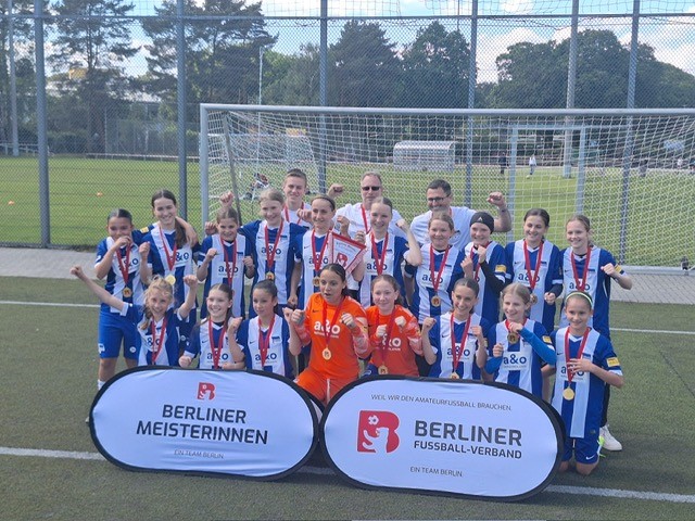 Vor einem Fußballtor auf einem Rasenplatz steht eine Gruppe junger Fußballspielerinnen in blau-weiß gestreiften Trikots. Viele von ihnen tragen Medaillen um den Hals. Vor der Gruppe liegen zwei Banner: Das linke zeigt die Aufschrift „BERLINER MEISTERINNEN“ mit dem Zusatz „Ein Team Berlin“. Das rechte Banner trägt die Aufschrift „BERLINER FUSSBALL-VERBAND“ sowie darunter „WIR SIND DER AMATEURFUSSBALL-BRANDENBURG“ und ebenfalls „Ein Team Berlin“.