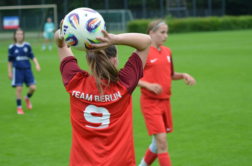 Das Bild zeigt eine dynamische Szene aus einem Fußballspiel auf einem gepflegten grünen Spielfeld. Im Vordergrund steht eine Spielerin in einem roten Trikot mit der Aufschrift „TEAM BERLIN“ und der Nummer 9. Sie bereitet sich darauf vor, den Ball einzuwerfen, hält ihn über dem Kopf und hat beide Arme nach hinten gestreckt. Im Hintergrund sind weitere Spielerinnen zu sehen – eine trägt ein blaues Trikot, eine andere ebenfalls ein rotes. Hinter dem Spielfeld sind Tore sowie einige Zuschauer oder Betreuer erkennbar.