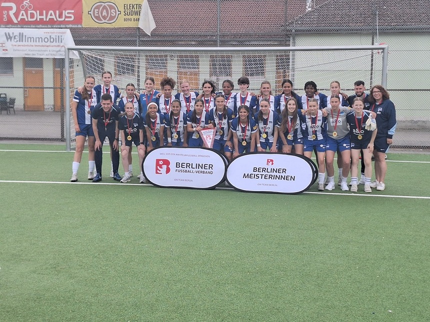 Auf einem Fußballfeld steht eine Gruppe von Personen, vermutlich ein Frauenteam, vor einem Tor und posiert für ein gemeinsames Foto. Die Teammitglieder tragen Medaillen und halten Pokale in den Händen – ein Hinweis auf einen gewonnenen Titel. Vor der Gruppe stehen zwei Schilder mit der Aufschrift „BERLINER FUSSBALL-VERBAND“ und „BERLINER MEISTERINNEN“.