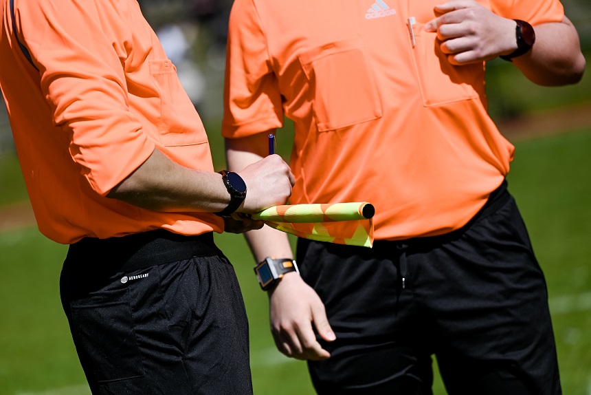Zwei Schiedsrichter in orangefarbenen Trikots stehen auf einem Sportplatz. Einer hält eine gelb-grüne Fahne und einen Stift, der andere zeigt mit dem Finger auf etwas. Beide tragen schwarze Hosen und Armbanduhren. Foto: Sandra Ritschel