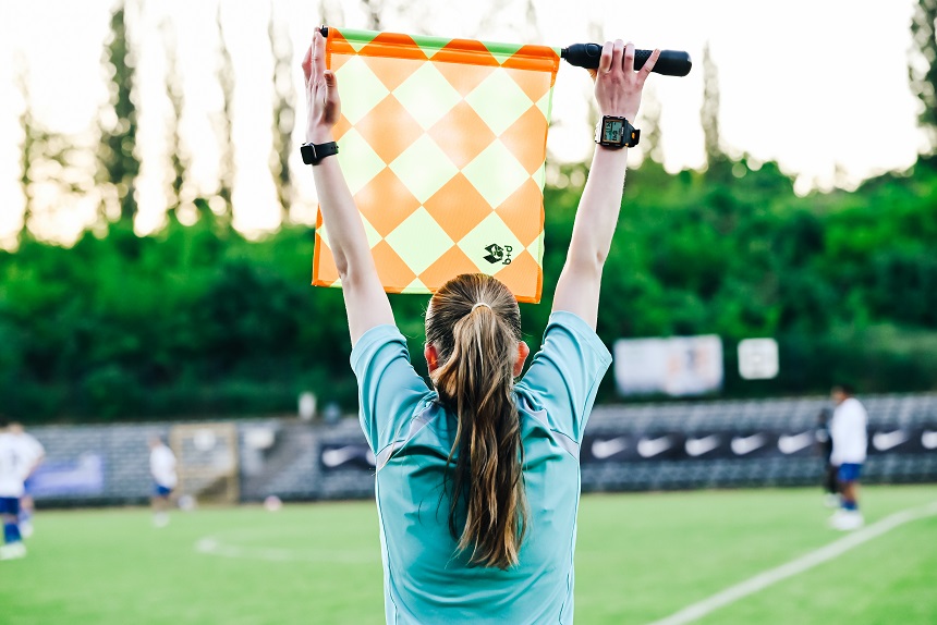 Eine Fußball-Schiedsrichterin steht am Spielfeldrand und hält eine orange-gelb karierte Fahne hoch, um eine Entscheidung anzuzeigen. Im Hintergrund sind Spieler, Bäume und Werbetafeln zu sehen. Foto: Sandra Ritschel