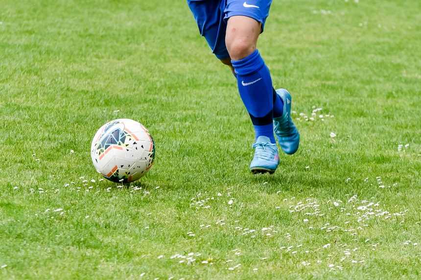 Auf dem Foto ist eine Person zu sehen, die auf einem grünen, mit kleinen weißen Blumen übersäten Rasen läuft. Die Person trägt ein komplettes blaues Fußball-Outfit: ein blaues Trikot, blaue Shorts, blaue Stutzen und blaue Fußballschuhe. Die Person ist gerade dabei, einen weißen Fußball mit schwarzen und orangefarbenen Mustern zu kicken. Der Moment wirkt dynamisch – es scheint, als sei die Person mitten im Spiel oder Training.