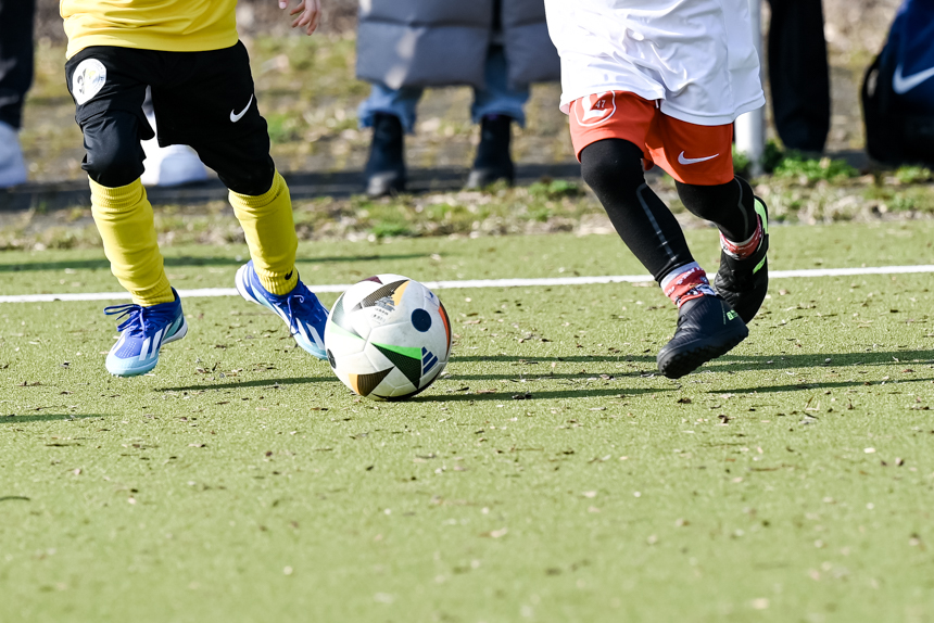 Nahaufnahme von zwei Kindern beim Fußballspiel auf einem Rasenplatz. Ein Kind trägt ein gelbes Trikot mit schwarzen Shorts und gelben Stutzen, das andere ein weißes Trikot mit roten Shorts und schwarzen Leggings. Beide sind in Bewegung und nahe am Ball. Im Hintergrund sind Teile des Spielfelds und Zuschauer am Spielfeldrand zu sehen.