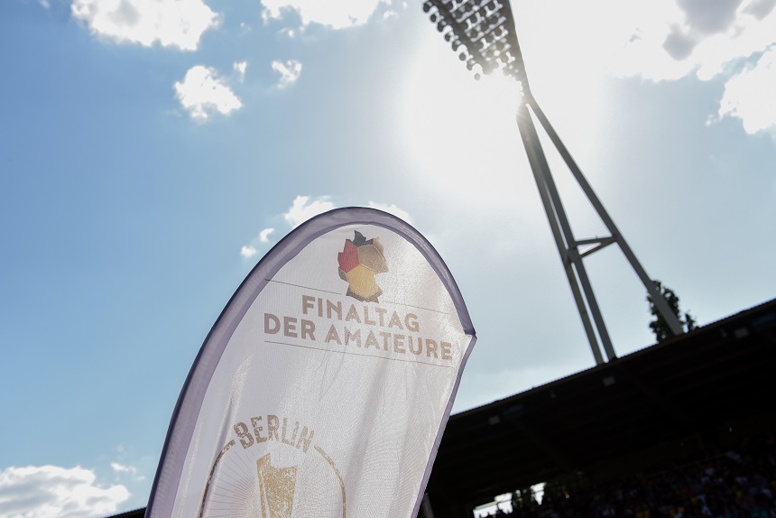 Das Bild zeigt eine Flagge mit der Aufschrift "FINALTAG DER AMATEURE" und "BERLIN". Im Hintergrund ist ein Stadion mit einem großen Flutlichtmast und einem teilweise bewölkten Himmel zu sehen.