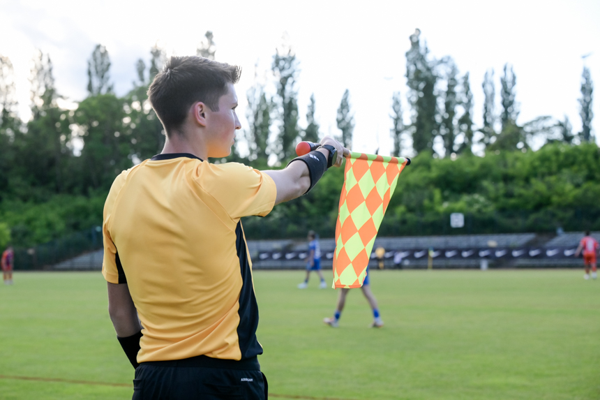 Auf dem Foto ist ein Schiedsrichterassistent auf einem Fußballfeld zu sehen. Er steht am Spielfeldrand und trägt ein gelbes Trikot mit schwarzen Details. In seiner rechten Hand hält er eine gelb-rot karierte Fahne, die typisch für Schiedsrichterassistenten ist. Im Hintergrund erkennt man mehrere Bäume, die das Spielfeld begrenzen. Auf dem Rasen befinden sich außerdem einige Fußballspieler – sie tragen blaue und rote Trikots. Die Szene spielt sich im Freien ab, vermutlich während eines Fußballspiels bei Tageslicht.