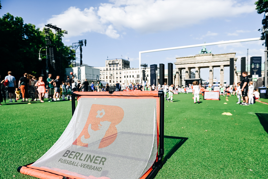 Auf einem temporären Fußballfeld mit grünem Kunstrasen vor dem Brandenburger Tor in Berlin steht im Vordergrund ein kleines Tor mit dem Logo des Berliner Fußball-Verbands. Im Hintergrund sind zahlreiche Menschen, darunter viele Kinder in bunter Sportkleidung, die sich aktiv an einem Sportevent beteiligen. Der Himmel ist blau mit vereinzelten weißen Wolken. FOTO: Sandra Ritschel
