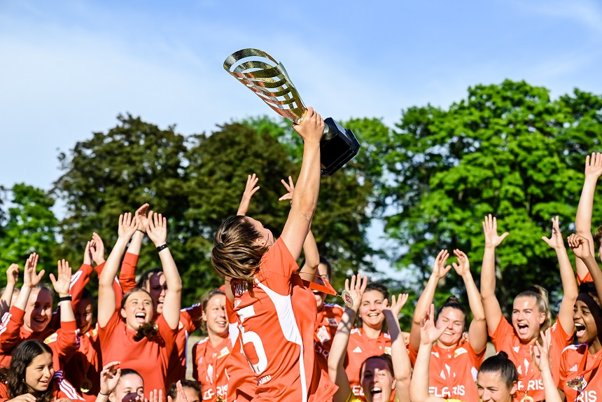 Eine Gruppe von Spielerinnen in roten Trikots feiert und hebt die Hände. Eine Person in der Mitte, mit der Nummer 15 auf dem Rücken, hält einen großen Pokal hoch. Im Hintergrund sind Bäume und ein blauer Himmel zu sehen. Foto: Sandra Ritschel