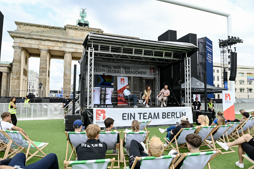 Das Foto zeigt eine öffentliche Veranstaltung vor dem Brandenburger Tor in Berlin. Im Vordergrund sitzen mehrere Menschen entspannt auf bunten Liegestühlen. Sie blicken auf eine Bühne, auf der drei Personen sitzen und offenbar an einer Diskussion oder einem Interview teilnehmen. Die Bühne ist mit Bannern geschmückt. Auf dem oberen Banner steht in großen Buchstaben: „WEIL WIR DEN AMATEURFUSSBALL BRAUCHEN!“. Darunter hängt ein weiteres Banner mit der Aufschrift: „EIN TEAM BERLIN“. Links neben der Bühne steht ein großes Schild mit dem Logo des Berliner Fußball-Verbands.