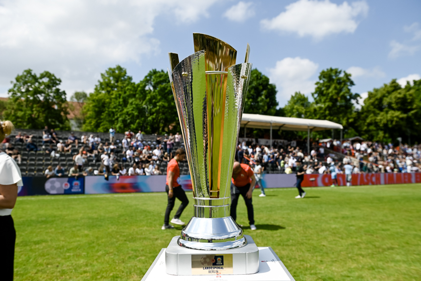 Ein großer, glänzender silberner Pokal steht auf einem weißen Sockel. Der Pokal hat ein kunstvolles Design mit mehreren vertikalen Elementen und einem breiten Sockel. Im Hintergrund ist ein Sportfeld mit grünem Rasen und Zuschauern auf den Tribünen zu sehen. Einige Menschen stehen oder gehen auf dem Feld, und in der Ferne sind Bäume unter einem teilweise bewölkten Himmel zu erkennen. Foto: Sandra Ritschel
