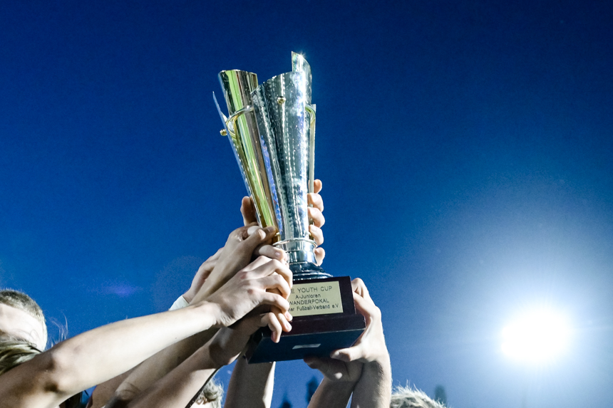 Mehrere Hände halten einen großen Pokal vor einem klaren blauen Himmel hoch. Der Pokal hat eine glänzende, metallische Oberfläche und ist mit Text graviert. Am Sockel des Pokals befindet sich eine Plakette mit der Aufschrift: "YOUTH CUP CHAMPIONSHIP WINNERS". Foto: Sandra Ritschel