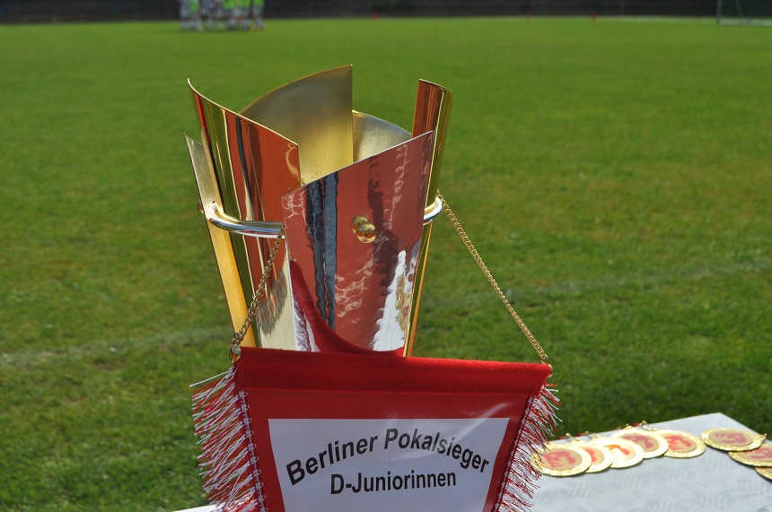 Ein goldener Pokal mit einem rot-weißen Banner daran. Auf dem Banner steht: "Berliner Pokalsieger D-Juniorinnen." Der Pokal steht auf einem Tisch, und im Hintergrund ist ein grasbewachsenes Feld zu sehen. Foto: BFV