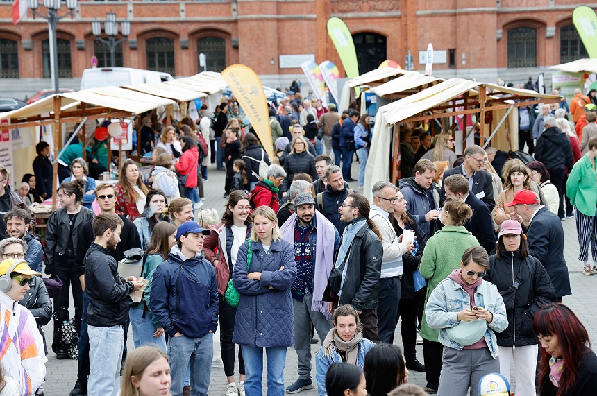 Das Foto zeigt eine belebte Szene auf einem offenen Markt oder Straßenfest. Zahlreiche Menschen bewegen sich zwischen mehreren Verkaufsständen, die aus Holzrahmen bestehen und mit Stoffdächern überspannt sind. Einige Personen unterhalten sich, andere betrachten die ausgestellten Waren oder benutzen ihre Mobiltelefone. Die Menschen sind unterschiedlich gekleidet, was auf eine vielfältige Besuchergruppe hinweist.  Im Hintergrund steht ein großes Gebäude aus roten Ziegelsteinen mit mehreren hohen, rundbogigen Fenstern und Türen. Über dem Markt sind verschiedene Banner und Schilder aufgehängt, die auf Angebote oder Veranstaltungen hinweisen könnten. Die Atmosphäre wirkt lebendig und einladend.