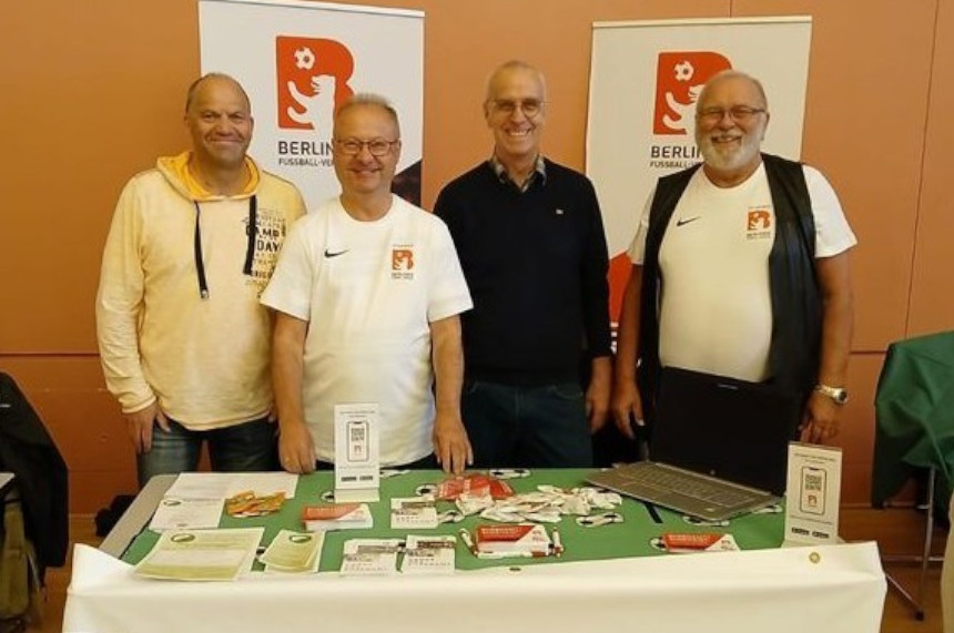 Das Foto zeigt vier Personen, die hinter einem rechteckigen Tisch stehen. Der Tisch ist mit verschiedenen Informationsmaterialien bedeckt, darunter Flyer, Broschüren, ein Laptop und weitere Werbematerialien. Die Personen stehen nebeneinander und blicken in Richtung Kamera. Einige von ihnen tragen T-Shirts mit dem Logo des Berliner Fußball-Verbands.