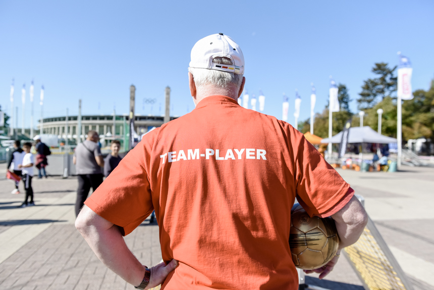 Das Foto zeigt eine Person von hinten. Sie trägt ein rotes T-Shirt mit der weißen Aufschrift „TEAM-PLAYER“. Die Person hat graue Haare und trägt eine weiße Baseballkappe. In der rechten Hand hält sie einen goldenen Fußball.  Im Hintergrund befinden sich mehrere Menschen, die sich auf einem offenen Platz bewegen. Es sind weiße Fahnen an Fahnenmasten zu sehen. Dahinter steht ein großes Gebäude, das wie ein Stadion aussieht. Der Himmel ist klar und blau.  Die Szene wirkt freundlich und offen, möglicherweise handelt es sich um eine Veranstaltung oder ein sportliches Treffen im Freien.