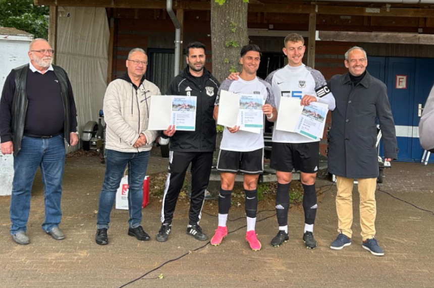 Auf dem Foto sind sechs Personen zu sehen, die nebeneinander stehen. Drei Personen in der Mitte tragen Sportkleidung und halten jeweils eine Urkunde in den Händen – sie scheinen für eine sportliche Leistung ausgezeichnet worden zu sein. Die anderen drei Personen stehen links und rechts neben den Sportlerinnen und tragen Alltagskleidung. Alle Personen stehen aufrecht und blicken in Richtung Kamera. Im Hintergrund ist ein Gebäude mit einer Holzwand zu erkennen, daneben steht ein Baum, der teilweise ins Bild ragt. Die Szene spielt sich im Freien ab, bei Tageslicht.