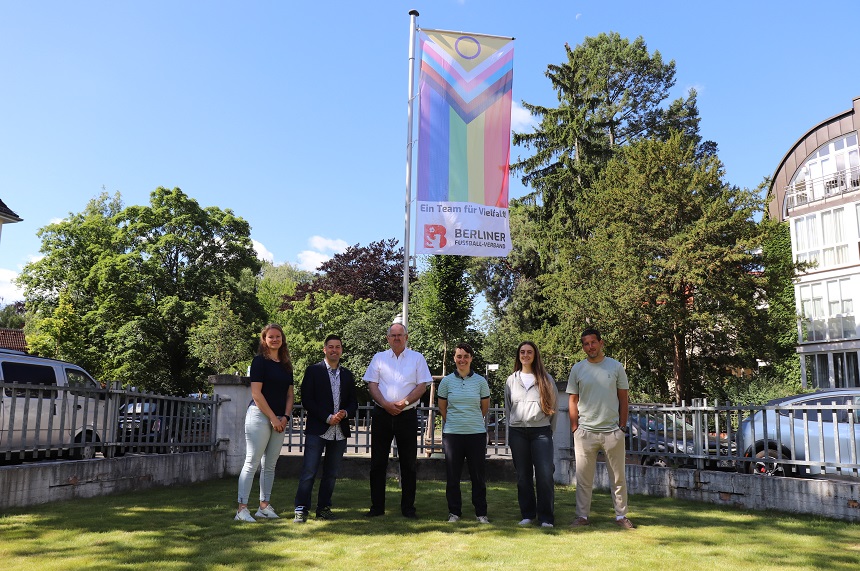 The photo shows six people standing together on a green lawn. They are standing next to each other in a loose formation and looking towards the camera. A large, colorful flag can be seen behind them. The banner reads "A team for diversity" in clearly legible lettering, accompanied by the logo of the Berlin Football Association. The colors of the flag are vibrant and symbolize diversity. Trees and buildings can be seen in the background, indicating an urban environment. The scene appears friendly and open. There are no visible barriers and the depiction emphasizes community and inclusion.