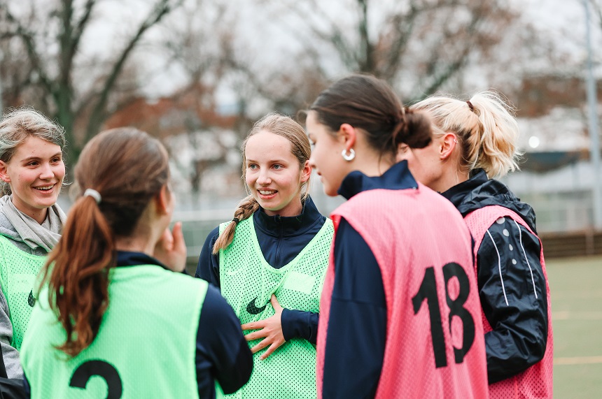 Das Foto zeigt sechs Personen auf einem Sportplatz. Alle tragen sportliche Kleidung und sind in zwei Teams aufgeteilt: Drei Personen tragen grüne Leibchen mit schwarzen Nummern, darunter eine mit der Nummer 3. Die anderen drei tragen pinke Leibchen. Die Körperhaltungen lassen darauf schließen, dass sie sich im Spiel oder Training befinden. Im Hintergrund erkennt man unscharf Bäume und einen Zaun, was auf eine Außenanlage hinweist. Die Szene wirkt dynamisch und vermittelt einen Eindruck von Teamaktivität und Bewegung.