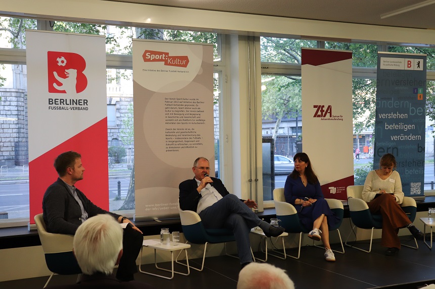 Das Bild zeigt eine Podiumsdiskussion mit vier Personen, die auf Stühlen vor Bannern sitzen. Die Banner enthalten Logos und Texte von verschiedenen Organisationen, darunter "Berliner Fußball-Verband," "Sport und Kultur," und "ZfA." Die Personen sind in ein Gespräch vertieft, wobei eine Person ein Mikrofon hält. Bei dieser Person handelt es sich um Bernd Schultz. Links neben ihm sitzt Moderator Ronny Blaschke. Rechts neben ihm sitzt Keren Vogler und Daniela Wurbs.