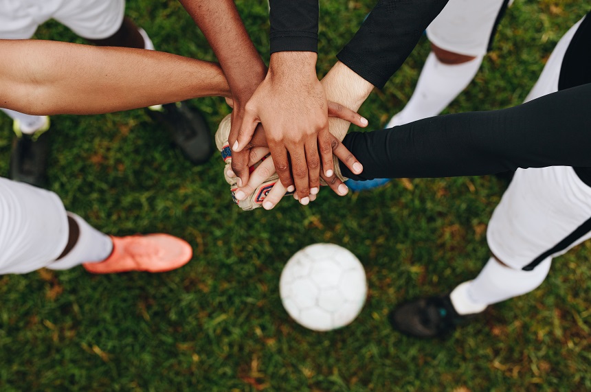 Das Foto zeigt eine Gruppe von fünf Personen, die im Kreis stehen und ihre Hände in der Mitte übereinandergelegt haben – ein Symbol für Teamgeist und Zusammenhalt. Alle Personen tragen Sportkleidung, vermutlich Fußballtrikots und -hosen. Die Hände unterscheiden sich in Hautfarbe, was auf eine vielfältige, inklusive Gruppe hinweist. Im Hintergrund liegt ein Fußball auf einem grünen Rasen, was die sportliche Atmosphäre unterstreicht. Die Perspektive des Bildes ist von oben aufgenommen, sodass der Fokus auf den Händen und dem Gemeinschaftsgefühl liegt.
