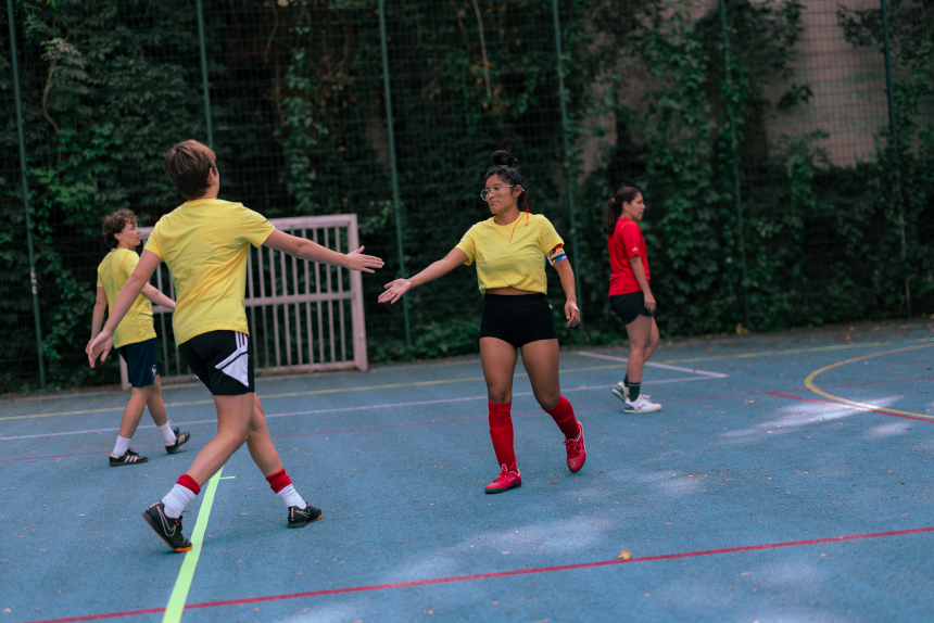 Mehrere Personen spielen auf einem Outdoor-Sportplatz; zwei Personen in gelben Trikots reichen sich die Hand, weitere stehen im Hintergrund. Foto: Loredana Zafisambondaoky