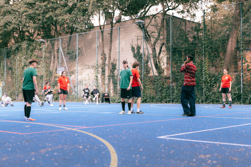 Mehrere Personen stehen auf einem blauen Outdoor-Sportplatz; weitere sitzen am Rand, Bäume und Zaun bilden den Hintergrund. Foto: Loredana Zafisambondaoky