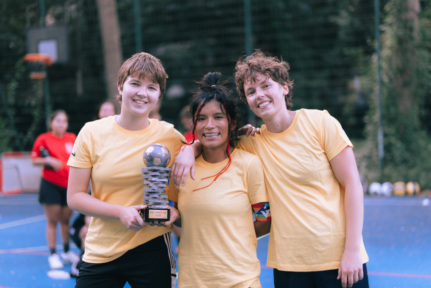 Drei Personen in gelben Trikots posieren auf einem Outdoor-Sportplatz mit einem Pokal; weitere Personen und Sportgeräte im Hintergrund. Foto: Loredana Zafisambondaoky