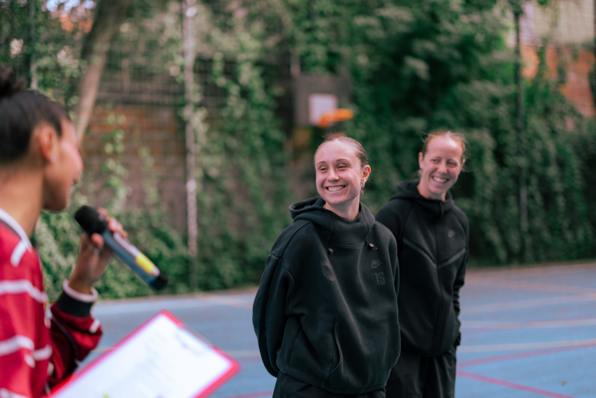 Person mit Mikrofon spricht auf einem Outdoor-Sportplatz zu zwei Personen in dunkler Sportkleidung; Bäume und Zaun im Hintergrund. Foto: Loredana Zafisambondaoky
